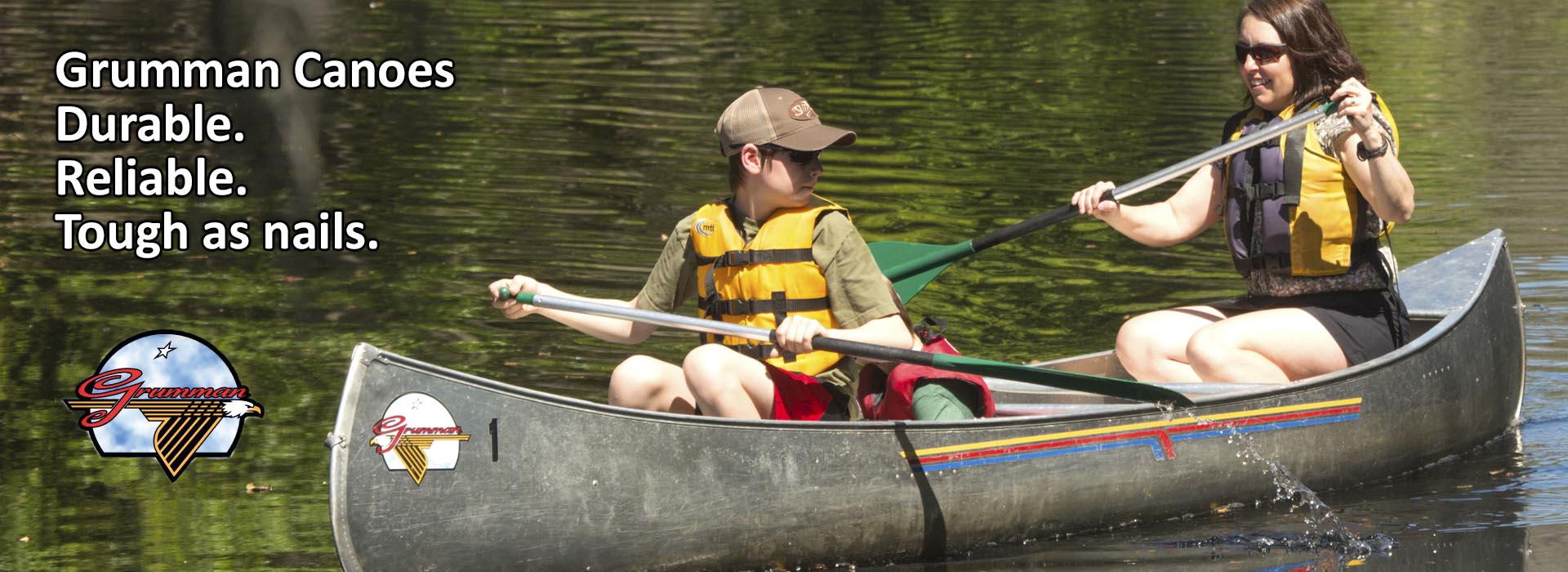 Grumman Canoes - Durable. Reliable. Tough as nails. Image shows a pre-teen white child with a hat and an adult white woman with brown shoulder length hair. Both are wearing yellow flotation vests, green canoe paddles, and are sitting in a grey Grumman canoe. The child is looking over their left shoulder back towards the adult. Water is trailing off of their canoe paddle indicating movement of the paddle