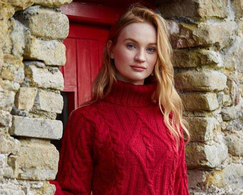 Shop Women's. Image description: A pale white woman with long red hair is standing in front of an old stone building with a red painted door. She is wearing a handcrafted Irish cabled sweater in a bright shade of red.