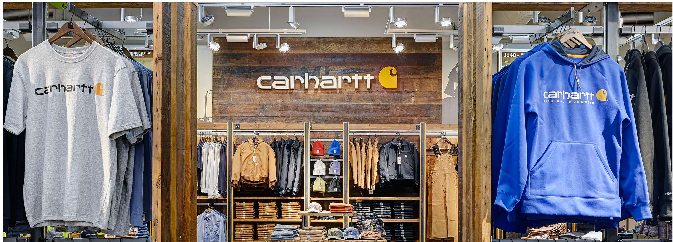 Shop Carhartt. Image description: A brand display of Carhartt clothing. In the foreground are display racks. On the left is a grey tee shirt with the Carhartt logo on the chest. On the right is a royal blue hooded sweatshirt with the Carhartt logo. In the background is a rustic wood wall with the Carhartt logo on a large sign over more displays of Carhartt work jackets, overalls, beanies, hats, and pants